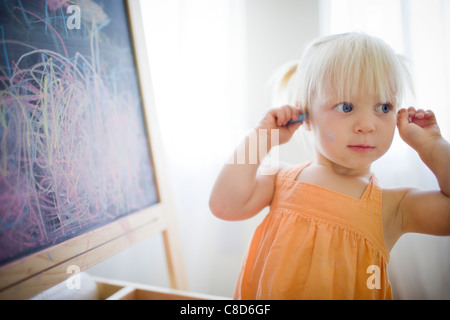 Toddler Girl at Chalkboard Putting Chalk in Her Ears Stock Photo