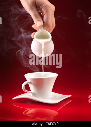 woman's hand pouring milk in white cup of coffee on red background Stock Photo