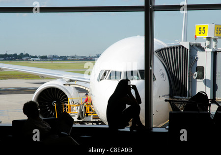A young woman on a mobile phone in silhouette in an airport departure lounge window with a plane behind Stock Photo