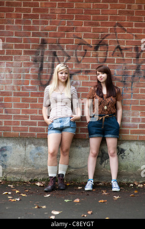 Two bored looking 16 17 year old teenage girls, standing in front of a brick wall UK Stock Photo