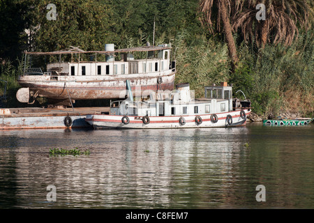 A section of Nile river bank with rusting old boat in dry dock with a single funnel boat moored in front reflected in the water Stock Photo