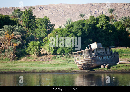 A section of Nile riverbank with abandoned derelict steel hull of ferry boat rusting away on the grassy edge, Egypt Stock Photo