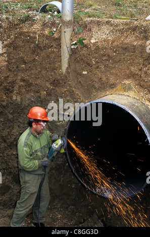 Slovakia. Man wearing an orange hard hat in an excavated hole working on a large gas pipeline using an angle grinder. Stock Photo