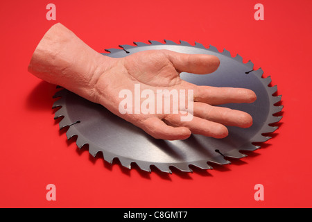 A fake human hand resting in the middle of a metal circular saw blade. Bright red background Stock Photo