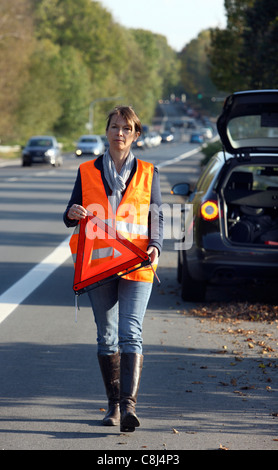 Car breakdown, on a highway, female driver, wearing a high visibility vest,  set up a warning triangle. Stock Photo