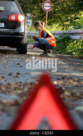 Car breakdown, flat tire. Woman changes a tire of a car on a highway, wearing a high visibility vest. Stock Photo