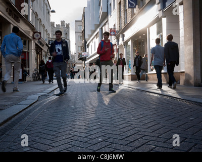 Shoppers in Cambridge walking in Market Street in backlit sunshine showing textures of road and long shadows. Stock Photo