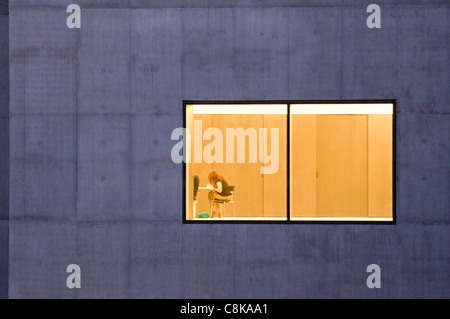 Grey concrete blocks & large window of Hepworth Gallery (lights on inside & woman sitting at table working) - Wakefield, West Yorkshire, England, UK. Stock Photo