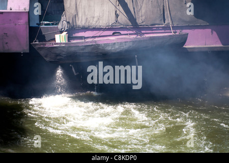 Small wooden boat tied to the stern of the smoking stern Nile of a Nile cruise boat navigating Esna lock Egypt Stock Photo