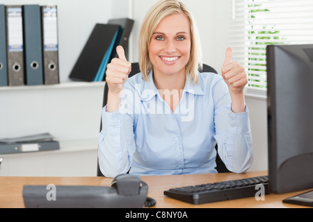 Young woman sitting behind desk with thumbs up Stock Photo