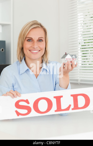 Gorgeous blonde businesswoman showing miniature house and SOLD sign looking into camera Stock Photo