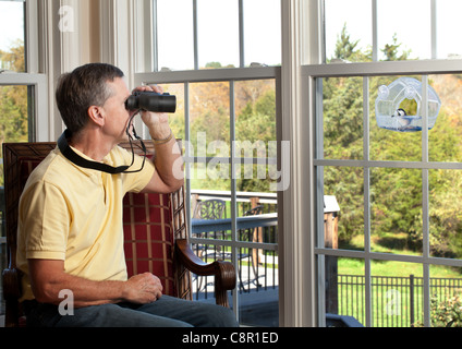 Senior man watching chickadee bird on birdfeeder through binoculars from chair Stock Photo