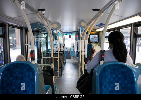Interior of public transport single deck bus & passengers seated inside back view woman watching security CCTV screen at front London England UK Stock Photo