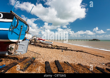 View along fishing boats on the shingle at Deal Beach in Kent, England, with the pier in the background on a summer's day, blue sky, white clouds. Stock Photo