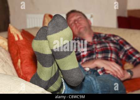 A tired millennial young man wearing striped socks jeans and a check shirt sleeping on a living room sofa. England UK Britain. Stock Photo