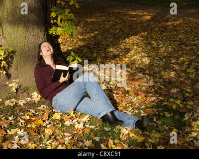 laughing out loud: a woman sitting benath a tree amidst fallen autumn leaves enjoying a funny book Stock Photo