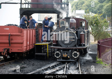 The engine driver of 2ft. Narrow Gauge steam locomotive double Fairlie 'Merddin Emrys' coals his engine from a coal wagon at Porthmadog station on the Ffestiniog Railway which runs between Porthmadog and Blaenau Ffestiniog, Snowdonia, North Wales, UK. Stock Photo