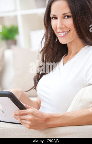 Beautiful young brunette woman at home sitting on sofa or settee using her tablet computer or iPad and smiling Stock Photo