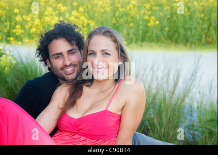 Young couple 20s sitting next to a pond in love happy smiling looking at camera sitting on grass she's leaning on boyfriend Stock Photo