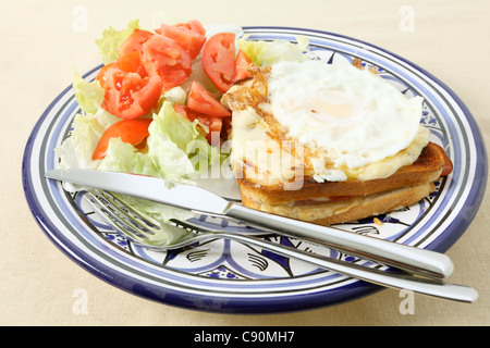 A croque madame - toasted cheese and ham sandwich topped with bechamel sauce and a fried egg - with a tomato and lettuce salad Stock Photo