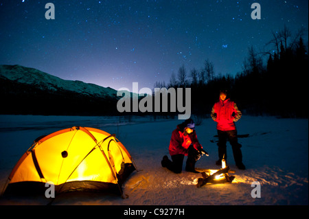 Two men warm themselves at a campfire while standing next to their lighted tent, Eklutna Lake in the Chugach State Park, Alaska Stock Photo