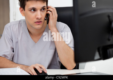 Doctor working on computer while talking on cell phone Stock Photo