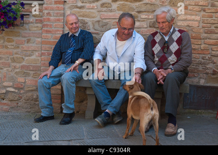 Three older adult Italian men sit on a bench midday petting a dog in Panicale, Italy. Stock Photo