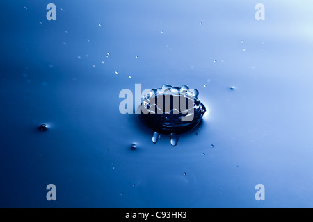 a drop of water hitting a pool of blue calm water creating a crater and causing a splash Stock Photo