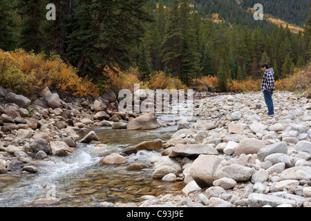 Asian man standing on rocky river bed enjoying nature, watching creek water flow by in autumn in Colorado Stock Photo
