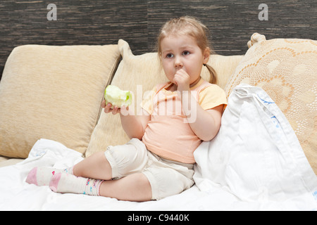 Little Russian girl sitting on sofa in domestic room and eating an apple Stock Photo