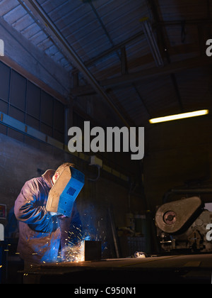 Manual worker in steel factory using welding mask, tools and machinery on metal. Vertical shape, side view, waist up Stock Photo