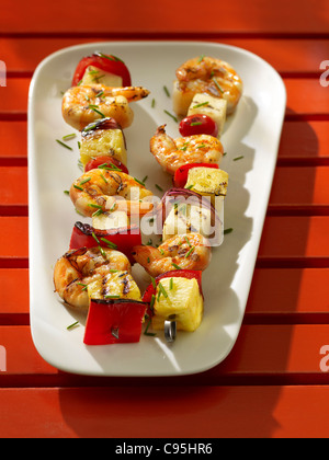 Grilled shrimp and vegetable brochettes with bell peppers, onions and pineapple on a white platter and red background Stock Photo