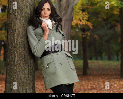 Fashionably dressed for autumn season, beautiful young woman in a green coat, leaning against a tree in a park. Stock Photo