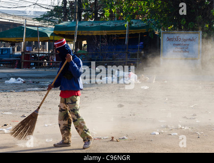 Young man with face mask and camouflage pants sweeping up rubbish with large straw broom after festival in northern Thailand Stock Photo
