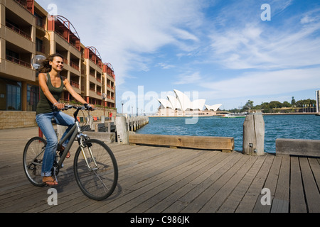 Woman riding bicycle along Sydney harbour with the Opera House in background. Sydney, New South Wales, Australia Stock Photo