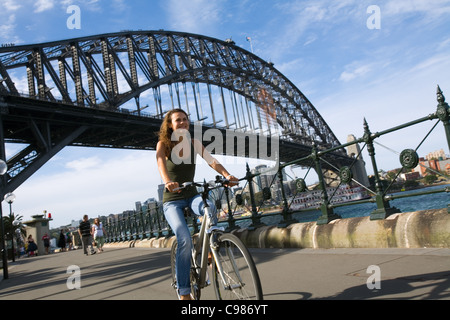 Woman riding bike along harbour with Sydney Harbour Bridge in background.  Sydney, New South Wales, Australia Stock Photo