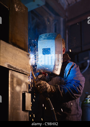 Manual worker in steel factory using welding mask, tools and machinery on metal. Vertical shape, side view, waist up Stock Photo