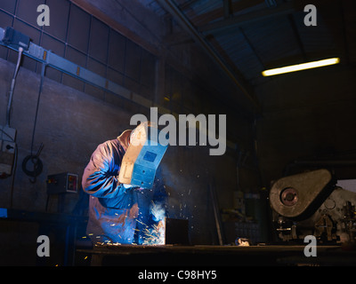 Manual worker in steel factory using welding mask, tools and machinery on metal. Horizontal shape, side view, waist up Stock Photo