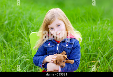 Blond kid girl with puppy pet dog sit in outdoor green grass Stock Photo