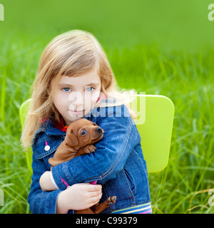 Blond kid girl with puppy pet dog sit in outdoor green grass Stock Photo