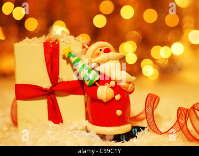 Holiday background with cute Santa Claus Christmas tree decorative ornament & gift box in snow over abstract defocus lights Stock Photo