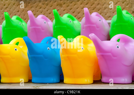 Colourful children's watering cans (in shape of elephants with trunk spouts) for sale & lined-up on 2 shelves - garden centre, Yorkshire, England, UK. Stock Photo