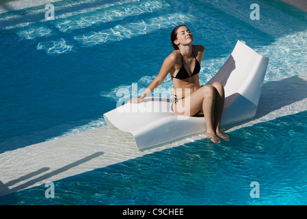 Young woman sitting on poolside deckchair dangling feet into water Stock Photo