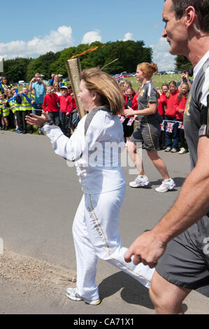 Olympic Torchbearer Janet Baker (carrying & holding flaming torch aloft) is watched by large crowd of well-wishers & accompanied by torch security team members in grey running kit. In the grounds of Harewood House, Yorkshire, England, UK. Tuesday 19th June 2012. Stock Photo