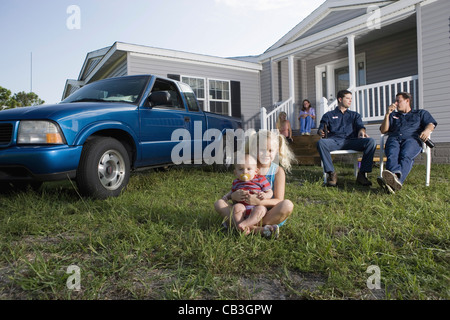 Young girl and a baby sitting on the front lawn of a trailer home, family in background Stock Photo