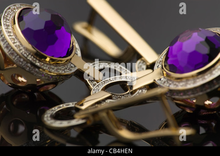 Macro snapshot of golden earrings with big purple gems on dark gray reflecting background. Small depth of field. Stock Photo