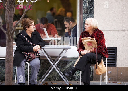 Two women sitting at sidewalk café table on Black Friday while man uses laptop inside window. Oak Park, Illinois. Stock Photo