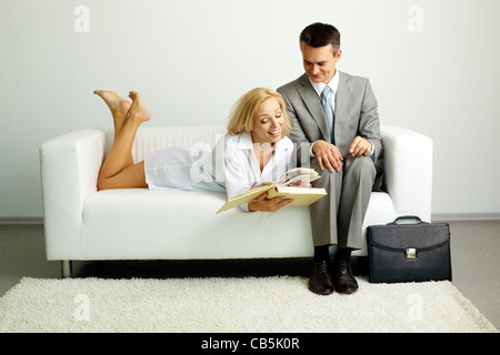 Photo of smiling man sitting on sofa with happy woman lying near by and both looking in book Stock Photo
