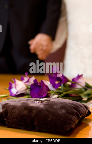 Wedding rings prepared before wedding ceremony on red cushion and bride and groom holding hands in background. Stock Photo