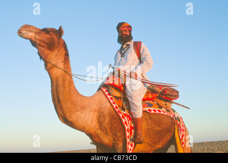 Mr Desert in traditional Rajput, Rajasthani dress carrying a sword, riding a camel on sand dunes near Jaisalmer, Rajasthan, India Stock Photo